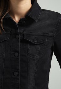 Chaqueta de mezclilla negra con cierre de botones, dos bolsillos en el pecho y una silueta ajustada. La tela tiene una textura suave y detalles de costura sutiles.