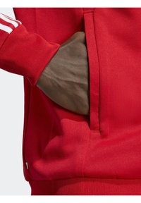 Červená sportovní mikina s klokáním kapsou a žebrovanými lemy. Má bílé pruhy na rukávech a hladkou texturu.