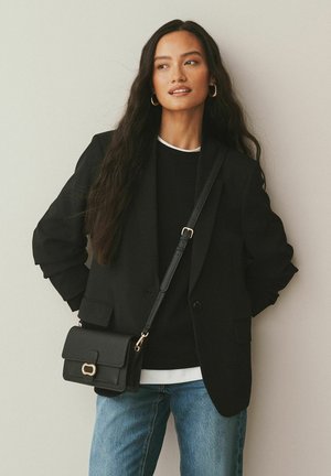 Schwarzer Blazer über einem schwarzen Pullover, kombiniert mit hellblauen Jeans. Eine kleine schwarze Tasche mit einem goldenen Akzent hängt an einem Riemen. Heller Hintergrund.