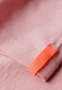 Helles rosa Gewebe mit glatter Textur, das ein auffälliges orangefarbenes Etikett mit erhabenen Text enthält, auf einer umgeschlagenen Kante und dem Aufdruck "SUPERDRY".