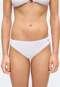 Žena v bílých žebrovaných bikinkách a košíčku bikinek se stejným vzorem a malým logem na středu přední části a boků na jednobarevném pozadí.