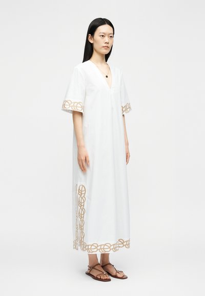 Femme debout portant une robe midi blanche avec de larges manches et une broderie circulaire beige sur les manches, l'ourlet et les côtés, associée à des sandales marron.