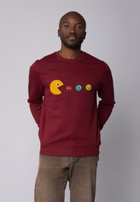 Sweatshirt bordeaux avec un graphique de Pac-Man jaune et quatre visages émoticons colorés alignés. Tissu doux, col rond, design ajusté.