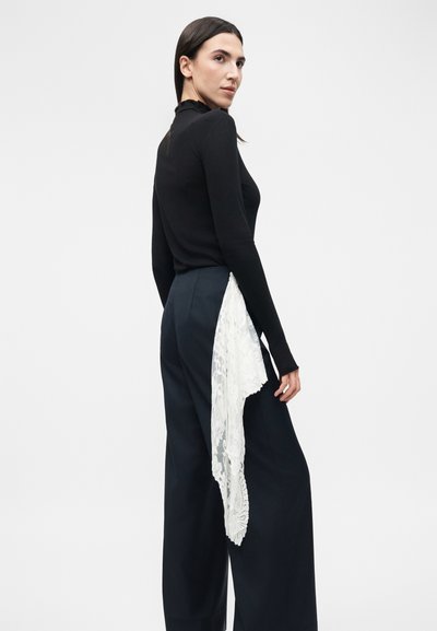 Γυναίκα που φοράει μαύρο μακρυμάνικο τοπ και σκούρο μπλε παντελόνι με φαρδιά πόδια, με λευκή δαντελένια λεπτομέρεια να κρέμεται από τη μέση.