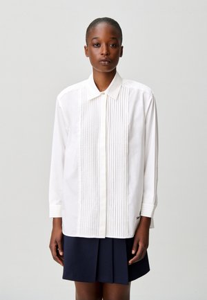 Una persona indossa una camicia bianca a maniche lunghe con pieghe verticali sul davanti e una gonna plissettata scura, in piedi davanti a uno sfondo semplice.