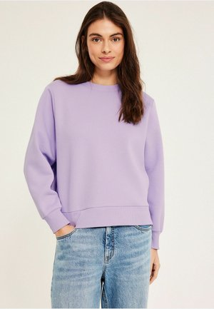 Vrouw met lang bruin haar, die een lavendelkleurige trui en lichtblauwe jeans draagt, staand tegen een effen lichte achtergrond.