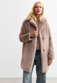 Cappotto in finta pelliccia beige chiaro con colletto a rever, due bottoni e tasche laterali. Aspetto testurizzato, abbinato a un top in maglia beige e jeans larghi.