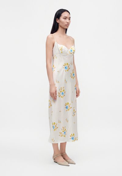 Jeune femme portant une robe midi blanche sans manches avec des imprimés floraux bleus et jaunes et des ballerines beiges pointues, debout devant un fond uni.