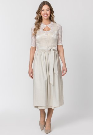 Frau trägt ein cremefarbenes traditionelles Kleid mit Spitzenärmeln, geknöpftem Mieder und gebundenem Taillenband, lächelnd vor einem einfarbigen Hintergrund.