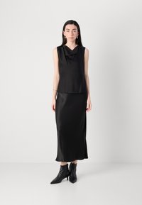 Svart sateng ermeløs topp med cowl-hals, kombinert med et matchende langt skjørt. Modellen har på seg spisse svarte ankelboots. Minimalistisk design.