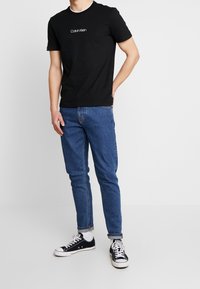 Černé bavlněné tričko s bílým logem Calvin Klein, doplněné modrými džínami a černými plátěnými teniskami, postavené před jednobarevným pozadím.