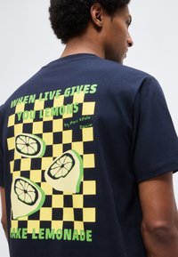 Άνδρας που φοράει μπλε ναυτικό t-shirt με μοτίβο κίτρινου καρό, τρεις φέτες λεμονιού και πράσινο κείμενο που γράφει «Όταν η ζωή σου δίνει λεμόνια, φτιάξε λεμονάδα.»