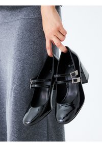 Zapatos de charol negro con punta cuadrada, que presentan doble correa y hebillas decorativas, descansando en una mano sobre un fondo textil gris.