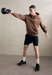 Barna kapucnis pulóver elülső zsebbel, fekete sportnadrág, fekete kettlebell a kezében, fekete sportcipő és fehér zokni semleges háttér előtt.