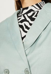 Vaaleansininen bleiseri, jossa on teksturoidu pinta, kaksirivinen malli ja vihreät napit, yhdistetty mustavalkoiseen seeprakuosiseen toppiin.