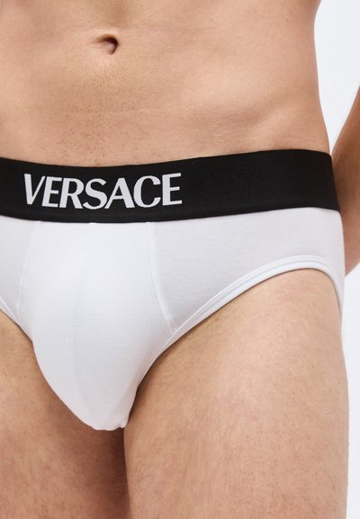 Homme portant un slip blanc avec une ceinture noire arborant le nom de la marque "Versace" en lettres blanches. Torse et hauts des cuisses visibles.