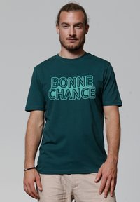 T-shirt en coton teal avec le texte "BONNE CHANCE" en police gras et en contour. Encolure ronde et manches courtes, avec un design minimaliste.