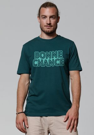 Teal katoenen T-shirt met de tekst "BONNE CHANCE" in vetgedrukte, omrande letters. Ronde hals en korte mouwen, met een minimalistisch ontwerp.