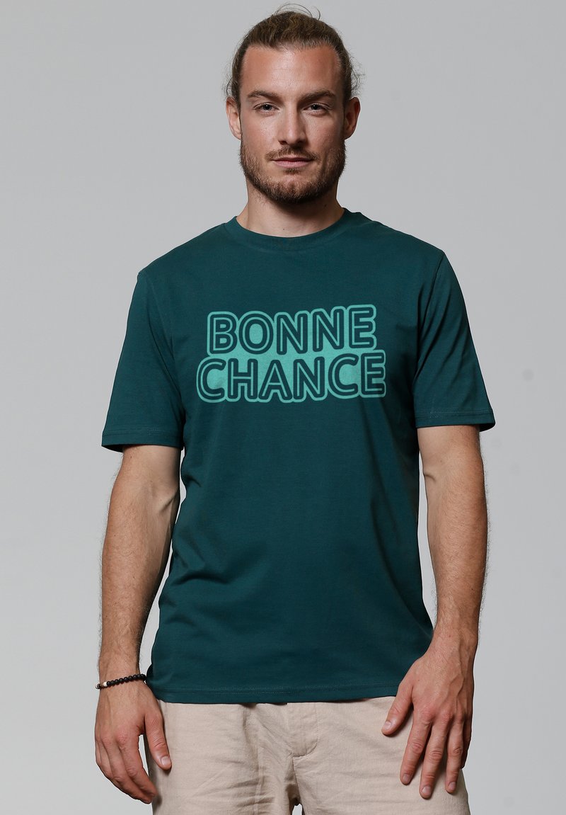 T-shirt en coton teal avec le texte "BONNE CHANCE" en police gras et en contour. Encolure ronde et manches courtes, avec un design minimaliste.