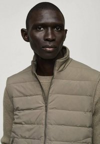 Vesta puffer bej cu guler înalt și față cu fermoar, având secțiuni matlasate și un design ajustat, purtată peste un top texturat bej.
