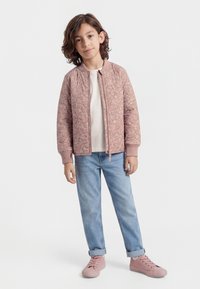 Kind mit schulterlangem braunem Haar, trägt eine rosa gemusterte Jacke mit Reißverschluss, ein weißes Hemd, hellblaue Jeans und rosa Turnschuhe, steht entspannt.