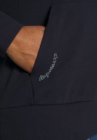 Sweat-shirt bleu marine foncé avec poche, main à l'intérieur et petit texte blanc brodé « Ragwear » avec détail en forme de cœur près du bord de la poche.