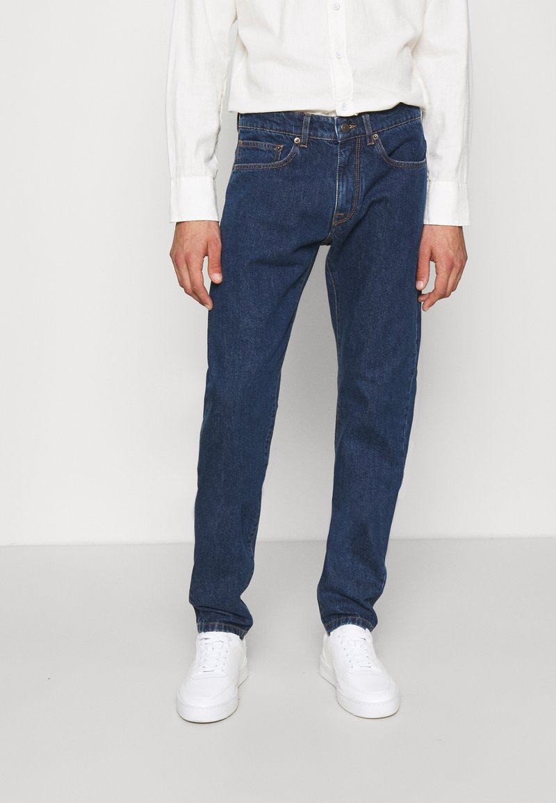 Homme portant un jean bleu foncé, une chemise blanche à boutons et des baskets blanches, debout devant un fond blanc uni.