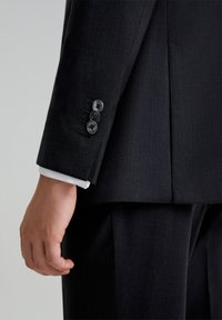 Chaqueta de traje negra ajustada con un primer plano de un puño que presenta tres botones negros y un puño de camisa blanca asomando. Textura suave.