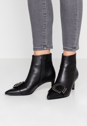 Bottines en cuir noir à bout pointu avec zips latéraux et boucles décoratives, portées avec un jean gris ajusté.