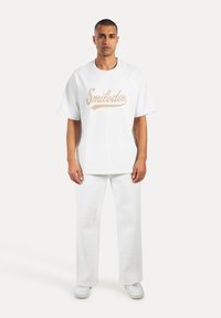 Hvid oversized T-shirt med beige "Smilodox" tekst, parret med hvide brede bukser. Materialet ser blødt ud og har en glat tekstur.