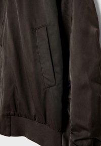 Crna bomber jakna s glatkom tkaninom, rebrastim donjim rubom i bočnom džepom s preklopom, koja prikazuje jednostavan, moderan dizajn.