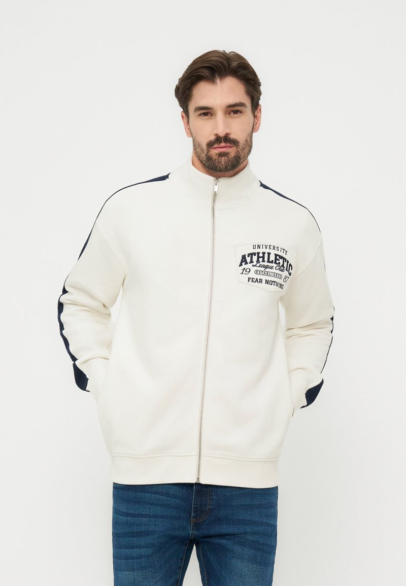 Άνδρας με μαύρα μαλλιά και γενειάδα, φοράει λευκό μπουφάν με φερμουάρ και μπλε ρίγες στα μανίκια, με την επιγραφή "University Athletic" στο στήθος, ποζάροντας με τα χέρια στις τσέπες.