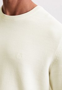Nahaufnahme einer Person, die einen strukturierten cremefarbenen Strickpullover mit rundem Ausschnitt und dezentem gestickten Logo auf der Brust trägt.