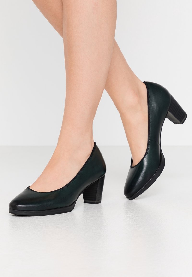 Legs wearing dark green leather low block heel shoes, standing on a smooth white floor, one foot slightly raised backward.