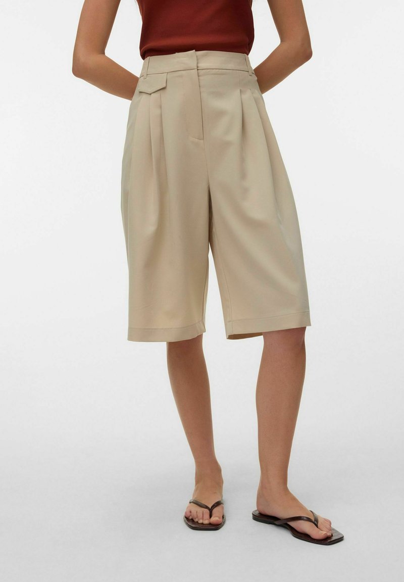 Femme portant un short beige coupé au genou avec des plis, associé à des tongs marron et un haut sans manches rouge foncé.
