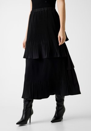 Femme portant une jupe midi noire plissée et superposée, associée à des bottines noires à talons aiguilles et à bouts pointus.