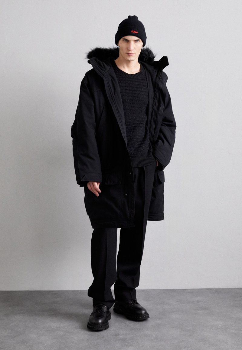 Schwarze Parka mit gefütterter Kapuze aus Fell, strukturierter schwarzer Pullover, schwarze Hose und schwarze Schuhe. Einfaches, modernes Design ohne sichtbare Logos.