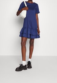 Mörkblå kortärmad klänning med lager på lager-volang i nederkant, tillverkad av mjukt tyg. Matchad med svarta skor med tjock sula och vita ankelsockor.