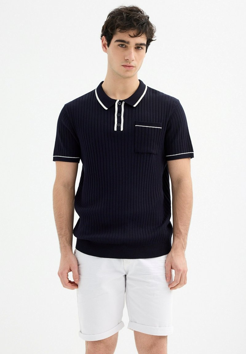 Polo de rayas en color navy con acentos blancos, tres botones y un bolsillo en el pecho, combinado con pantalones cortos blancos enrollados. Textura suave, corte ajustado.