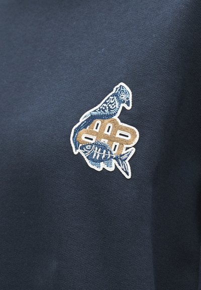 Tissu bleu avec un patch brodé montrant un oiseau bleu, un squelette de poisson stylisé et des formes géométriques brunes sur fond foncé.
