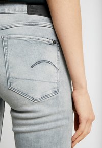 Jeans de mezclilla de corte slim en un azul claro, con un bolsillo trasero que presenta un detalle de costura curva y una etiqueta negra con el logo cerca de la cinturilla.