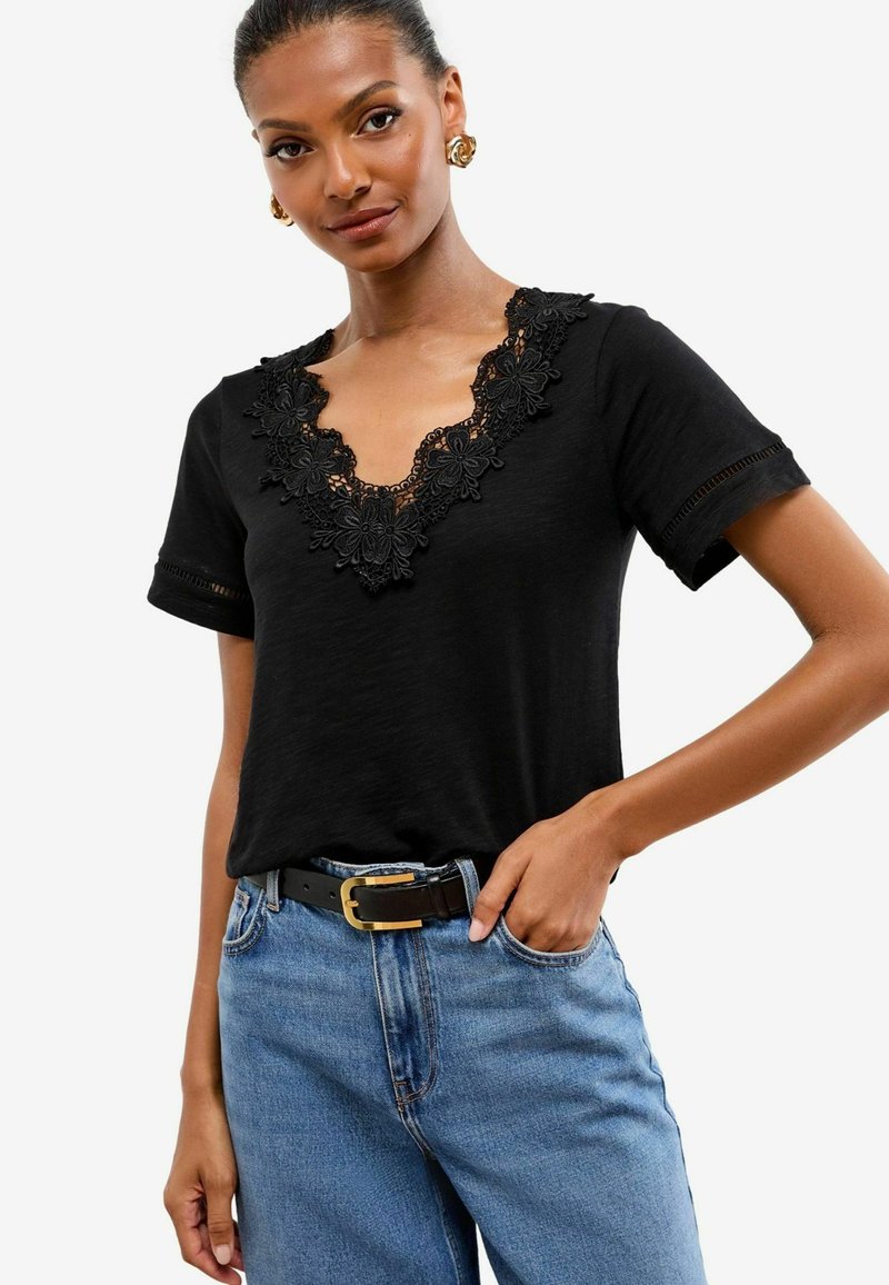 Melnā īsām piedurknēm T-kreklā ar dziļu V veida kakla izgriezumu, kas rotāts ar smalku melnu mežģīni. Savienots ar ziliem džinsiem un melnu jostu.