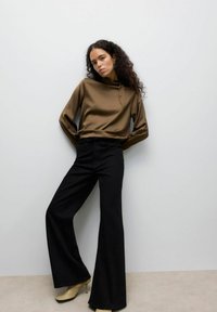 Haut long à manches en satin marron avec un col haut, associé à un pantalon noir évasé et des talons clairs, sur un fond neutre.