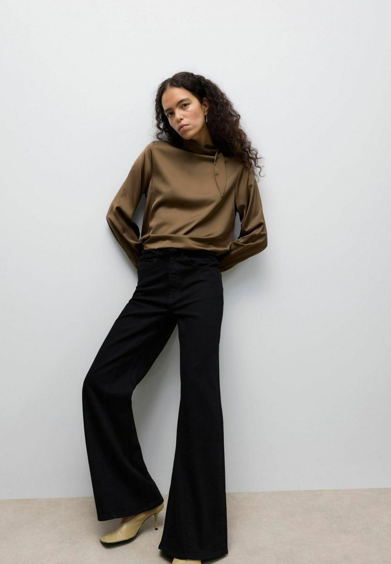 Haut long à manches en satin marron avec un col haut, associé à un pantalon noir évasé et des talons clairs, sur un fond neutre.