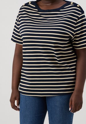 Mujer que lleva una camiseta de manga corta con rayas horizontales en azul marino y crema, con botones dorados en las costuras de los hombros, combinada con vaqueros azules.