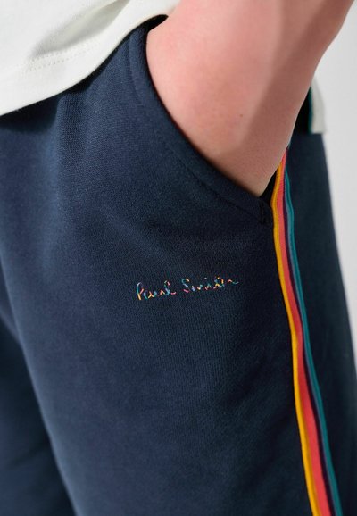 Main d'une personne dans la poche d'un pantalon bleu marine avec des bandes latérales multicolores et un petit logo brodé multicolore près de la poche.