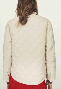 Prošívaná krémová bunda s výrazným límcem a knoflíky potaženými tkaninou, s dlouhými rukávy a hladkou texturou, nošená přes červený oděv.