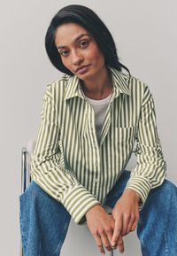 Camicia a righe verdi e bianche con bottoni, maniche lunghe e taschino sul petto, abbinata a jeans di denim blu. Design semplice, vestibilità casual.