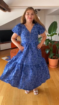 Μπλε φλοράλ μάξι φόρεμα με tiered φούστα, κοντά πλισέ μανίκια και V-λαιμόκοψη. Φοριέται με λευκά σανδάλια. Εσωτερικός χώρος με φυτά.