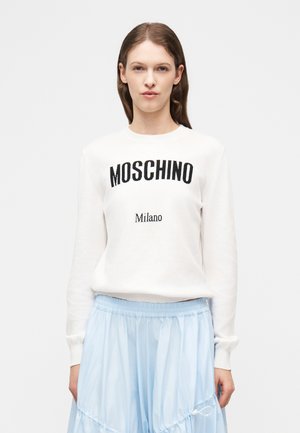 Femme portant un pull blanc avec le texte "MOSCHINO Milano" et une jupe bleu clair, debout devant un fond blanc uni.